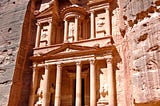 A pink-hued building in the Roman-Greek style with Corinthian columns is carved into a flat rockface