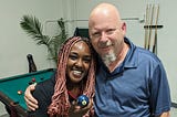 Partners Amani and Doug pose for a picture in front of a pool table while holding the 2 ball