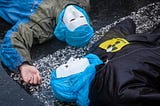 A photograph of two people lying on the ground, wearing white masks and with their heads wrapped in blue plastic.
