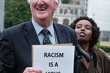 White man holding a sign smiling and a black woman standing beside him. The sign reads “Racism is a virus, we are the vaccine.”