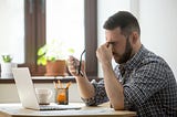 Photo of a stressed out man, sitting at a desk, in front of a laptop. He has his glasses in his right hand and, with his left, is squeezing the bridge of his nose. Photo courtesy of Freepik