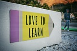 Picture of a white small wall with a sign on it in the shape of a pencil. On the sign it reads “love to learn”.