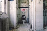 A white bathroom with dirty walls with a smiley face painted on the toilet lid