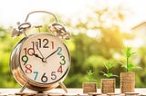 Image of a colorful analog alarm clock and three stacks of coins increasing in size with plant sprouts on them.