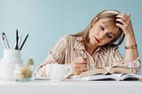 A thoughtful woman reads and writes at a desk with a blue wall behind her and a book open.