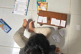 Seorang anak kecil duduk di lantai sedang belajar matematika dengan menggunakan potongan kertas bertuliskan angka. Di depannya terdapat meja kecil kayu dengan angka 1, 2, 6, 9, dan 4 yang ditempel berurutan. Di lantai juga terlihat buku latihan matematika anak, beberapa kertas angka lainnya, dan alat tulis. Suasana menunjukkan aktivitas belajar sambil bermain di rumah