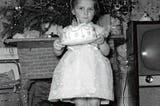 B&W photo of a girl holding a birthday cake in front of a fireplace with a Christmas tree ontop