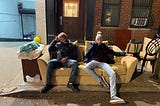 author (on left) and his friend (on right) sitting on a beige couch that is on a sidewalk. There is a chair and a cabinet on the sidewalk next to the couch