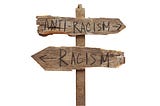 a rustic wooden signpost with an arrow pointing backwards to racism and forwards to anti-racism