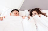 Two people in bed together with the sheets pulled up over their faces so that just their eyes are peeking out