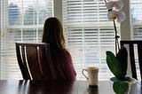 Photo of a woman looking out the window. Her coffee cup and an orchid are on the table behind her.