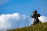 color photograph of a lone cross burial marker on crispy grass against a bright blue sky with cottony clouds