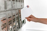 Picture of a hand placing a white stamped envelope with no writing on it into a bank of mailboxes.