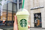 A hand holds up a green, frothy Starbucks beverage in front of an industrial-looking Starbucks. Maybe they’re about to Pay It Forward?