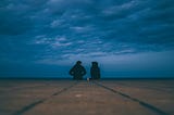 Two people seen from far away. They are sitting on the end of a pier, under a cloudy twilight sky