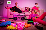 1980s-scene with two women splayed on couch, a boom box, mannequins and clothings strewn about