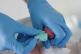 Two hands in blue medical gloves take a blood sample from a newborn’s heel.