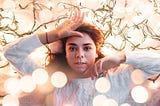 Woman with long dark hair wearing a white sweater lying in a pile of white Christmas lights. She is looking up at the camera.