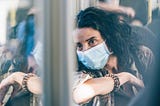 A woman in a surgical mask looks pensively out the window of a bus.