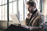 A white man in his 30s wearing a brown jacket and black turtleneck stares at his laptop as though pondering a dilemma.