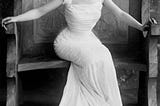 A promotional photograph from 1900 depicting British actress Ethel Warwick in a flowing white dress, looking into the camera while seated on an oversized chair.