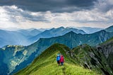 Two people walking a green mountain range with sunlit clouds cascading overhead.