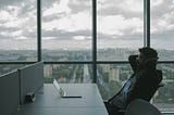 Man sitting and thinking at a desk in a corporate office with large windows and city scape in the background.