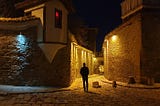 A man walks down a cobblestone road at night, buildings are on both sides of the narrow roadway.