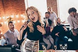 Pretty blonde woman in shiny shirt sings Karaoke infront of a crowd of friends.