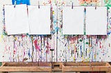 Four blank pages hang on paint covered easels in an elementary school classroom.