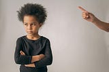 Young Black girl stands with her arms folded across her chest as a parent or guardian points a finger as they correct the child’s behavior.