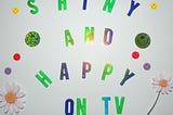 A poster with a white background and the words “Shiny and Happy on TV”, decorated with smiley faces and daisy flowers