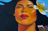 Chalk drawing of a woman’s face. The woman has long black hair, bright red lipstick, light brown skin, and a white flower in her hair. A box of pastel chalks is in the lower right corner of the photo.
