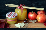 The makings of applesauce are arranged neatly on a wooden cutting board, including reddish-yellow apples, jars of chunky yellow sauce, a wooden spoon, with fall decor details like gingham ribbons around the spoon and one jar and as a pattern on the jar’s lid, two little wooden heart cut-outs, and a scrap of burlap.