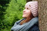 Photo of a young woman leaning against a tree trunk smiling and looking up.