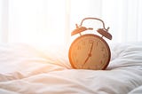 A clock sits on top of a white comforter with sun streaming in from the background