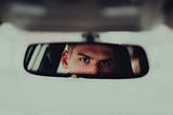 A young human with an intense look in their eyes looks at themselves in a car rearview mirror.