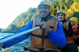 A man and two women paddle a kayak on a river. All three have big smiles and are  wearing yellow helmets and orange life vests. Tall green trees line the river on the right side of the photo.