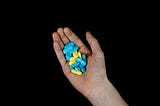 A person’s hand against a black backdrop, holding a mixture of blue and yellow pills.