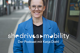 A woman stands smiling at a train platform, wearing a bright blue coat and glasses. Behind her is a modern train, suggesting a focus on public transportation. Overlaid text reads “She Drives Mobility — Der Podcast mit Katja Diehl.” A stylized waveform in the top right corner emphasizes the podcast theme. The image visually promotes a podcast about sustainable and inclusive mobility.