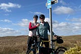 Two grey-bearded cyclists posing with their bikes by a National Byway sign. There is a blue sky dotted with white clouds behind them.