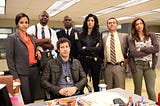 Three men and three women standing looking at the viewer and one man sitting looking sideways, in front of a desk in a police precinct.