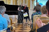 Two white women standing in front of a seated audience, one presenting an award to the other. A grand piano is visible in the background