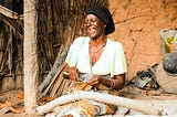 An elderly African woman sits outside a traditional mud-walled home, joyfully smiling and weaving natural fiber rope by hand. She wears a black headscarf, a light green blouse, patterned wrap skirt, and several bracelets. The setting is earthy and rustic, with palm fronds, a tree trunk, and household items around her, reflecting a rural environment and traditional craftsmanship.