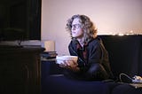 Person with light curly hair holding a popcorn bowl and illuminated by a TV screen they’re staring intently into