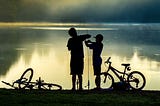 The silhouettes of two boys in front of a lagoon with their bicycles lying at their feet