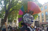 A head and shoulders of a punk with a rainbow-coloured mohawk surrounded by flowers and a bench set in a town square with trees and shops in the background and people milling around.