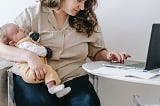 Woman holding baby in one arm while seated, looking at her laptop screen