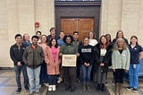 17 people pose outside a building with a brown door. Most wear jackets, and one holds a sign bearing the MICRO logo.