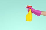 An arm in a pink plastic glove reaches into frame from the right side, holding a yellow spray bottle with a red top. The background is a light seafoam or turquoise.